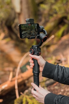 A person holds a smartphone mounted on a stabilizing gimbal in an outdoor setting. The smartphone screen displays a recording of a forest scene. The gimbal is black with red accents and features various controls. The person's hand is visible, wearing a dark jacket. The background consists of blurred natural elements, likely trees and foliage.