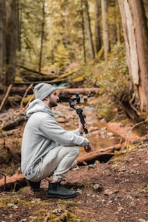 A person squats in a forest, holding a gimbal with a smartphone attached. They are wearing a gray hoodie, white pants, a beanie, and black boots. The surrounding area is full of tall trees, moss-covered logs, and a small stream running through the terrain.