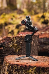 A modern camera stabilizer positioned on a tree stump in a forest setting. The stabilizer has a sleek design with black and red accents and is mounted on a small tripod. The background is blurred, highlighting the lush, moss-covered ground and trees.
