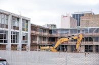 A construction site with partially demolished buildings and an excavator. The site is surrounded by a metal fence. The buildings appear old, with exposed rebar and boarded windows. In the background, high-rise buildings such as a hotel and office buildings are visible.