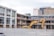 A construction site with partially demolished buildings and an excavator. The site is surrounded by a metal fence. The buildings appear old, with exposed rebar and boarded windows. In the background, high-rise buildings such as a hotel and office buildings are visible.