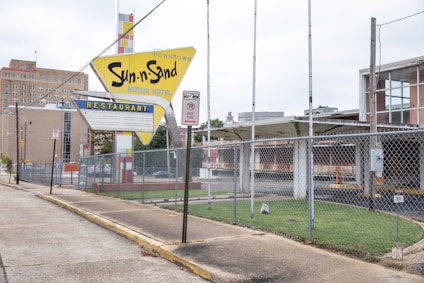 An aged and weathered sign for the Sun-n-Sand Motor Hotel stands in front of an old building. The sign has a mid-20th-century design and features a large arrow with bold yellow and blue colors. There's a chain-link fence in front of the vacant parking area, and a no parking sign is visible on a pole nearby. The surrounding area looks desolate with empty parking spaces and an overgrown lawn.
