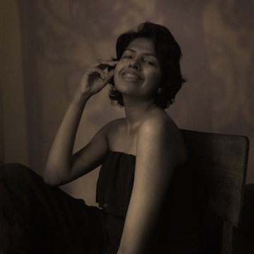 A person with short hair, wearing a strapless top and braces on their teeth, is seated on a chair with a gentle smile. The lighting creates a soft, sepia tone that adds a vintage feel to the portrait.