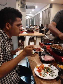 A man is seated at a wooden table inside a busy restaurant, eating from a plate filled with colorful vegetables and greens. There is a grill on the table with food being cooked, and various dishes and ingredients are spread around. The restaurant is bustling with other diners in the background and has a modern, casual ambiance.