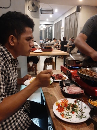 A man is seated at a wooden table inside a busy restaurant, eating from a plate filled with colorful vegetables and greens. There is a grill on the table with food being cooked, and various dishes and ingredients are spread around. The restaurant is bustling with other diners in the background and has a modern, casual ambiance.