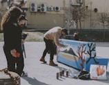 A person in casual clothing is painting a piano with a scenic design featuring mountains and trees, using spray paint. Nearby, two individuals are observing, one holding a red phone. The setting is an urban outdoor area with trees and a wall with air conditioning units in the background.