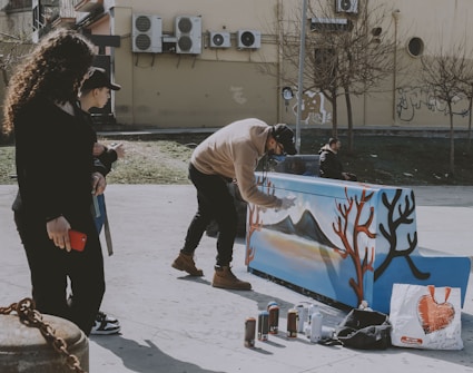 A person in casual clothing is painting a piano with a scenic design featuring mountains and trees, using spray paint. Nearby, two individuals are observing, one holding a red phone. The setting is an urban outdoor area with trees and a wall with air conditioning units in the background.