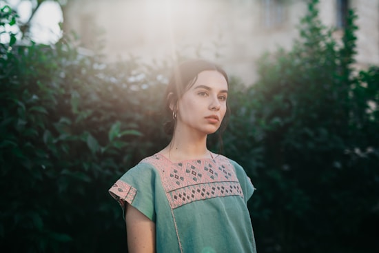 A young woman stands outdoors in front of lush green foliage. She wears a dress with intricate geometric embroidery in soft pink and green colors. Sunlight filters through the leaves, creating a soft, ethereal glow around her.