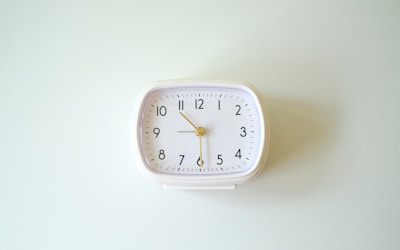 A white, rectangular analog clock with black numbers and a gold second hand, mounted against a light-colored wall. It displays the time as five minutes past ten.