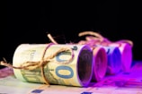 Several rolled bundles of euro banknotes are tied with twine, arranged in a row on a flat surface with a dark background. The lighting creates a gradient of purple and pink hues across the banknotes, adding a modern and abstract feel.