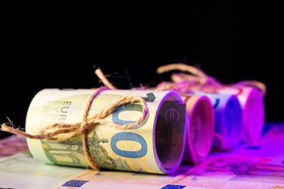 Several rolled bundles of euro banknotes are tied with twine, arranged in a row on a flat surface with a dark background. The lighting creates a gradient of purple and pink hues across the banknotes, adding a modern and abstract feel.