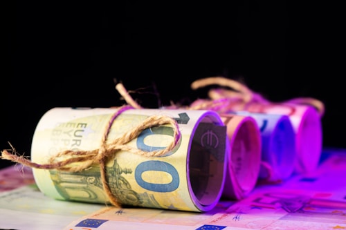 Several rolled bundles of euro banknotes are tied with twine, arranged in a row on a flat surface with a dark background. The lighting creates a gradient of purple and pink hues across the banknotes, adding a modern and abstract feel.