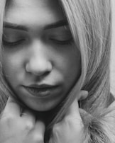 A close-up, black and white portrait of a person with eyes closed, suggesting introspection or contemplation. The individual's hands are gently positioned near the face, with long, light-colored hair framing the scene.