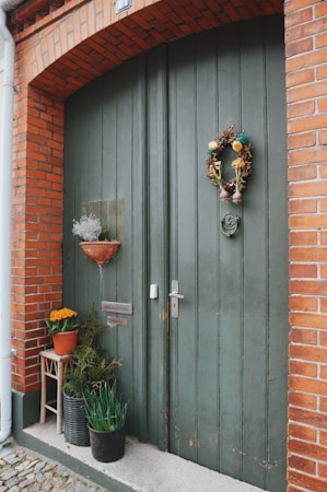 A large, rustic green wooden door is framed by red brickwork. A decorative wreath hangs in the center, adorned with dried plants and colorful ornaments. Potted plants are arranged beside the door, including vibrant yellow flowers, a potted evergreen, and another plant in a dark pot. A small, wooden stool supports one of the flower pots.