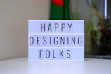 A lightbox displaying the words 'HAPPY DESIGNING FOLKS' is placed on a white surface. In the background, a blurred image of a flag with green and red colors is visible, along with some plants.