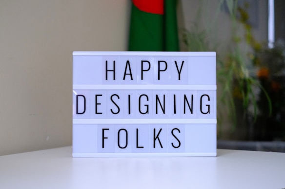 A lightbox displaying the words 'HAPPY DESIGNING FOLKS' is placed on a white surface. In the background, a blurred image of a flag with green and red colors is visible, along with some plants.