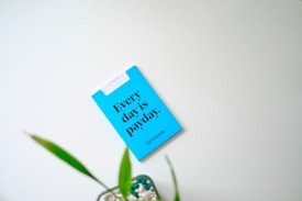A blue card with bold black text reading 'Every day is payday.' is affixed to a light-colored wall. Below the text, the name 'GoDaddy' is printed in smaller font. A potted plant with green leaves is partially visible at the bottom of the image.