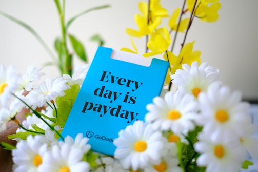 A bright blue card with bold black text saying 'Every day is payday' surrounded by a cluster of white daisies with yellow centers and a few yellow flowers against a blurred background.