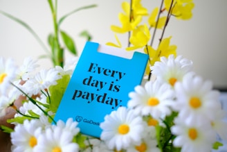 A bright blue card with bold black text saying 'Every day is payday' surrounded by a cluster of white daisies with yellow centers and a few yellow flowers against a blurred background.