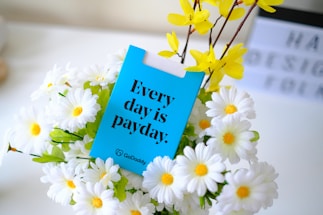 A vibrant arrangement of white daisies with yellow centers and yellow flowers is displayed, with a bright blue card placed among them. The card features bold black text reading 'Every day is payday', accompanied by a small logo in the bottom corner. In the background, there is a blurred white sign with indistinct letters in black.