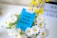 A vibrant arrangement of white daisies with yellow centers and yellow flowers is displayed, with a bright blue card placed among them. The card features bold black text reading 'Every day is payday', accompanied by a small logo in the bottom corner. In the background, there is a blurred white sign with indistinct letters in black.