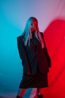 A person stands confidently, wearing a large dark suit against a backdrop with strong contrasting red and blue lighting. The lighting creates a dramatic and moody atmosphere, casting shadows and highlights on the subject's face and the large, oversized jacket.