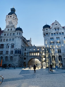 An architectural scene featuring a grand, historic stone building with tall towers and intricate details, under a clear blue sky. Sunlight creates patterns of shadows on the facade. Several people are walking and cycling through the cobblestone courtyard below.