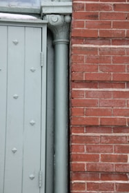 The image displays a section of a building exterior featuring a light green door with embossed diamond patterns beside a vertical downpipe or column. Adjacent to the door is a red brick wall with a clear, straight line of mortar.