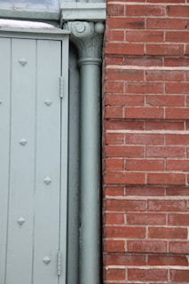 The image displays a section of a building exterior featuring a light green door with embossed diamond patterns beside a vertical downpipe or column. Adjacent to the door is a red brick wall with a clear, straight line of mortar.