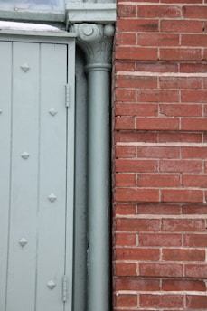The image displays a section of a building exterior featuring a light green door with embossed diamond patterns beside a vertical downpipe or column. Adjacent to the door is a red brick wall with a clear, straight line of mortar.