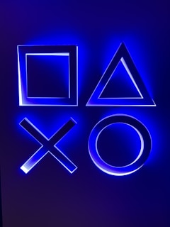 Illuminated geometric shapes including a square, triangle, cross, and circle, glowing with a blue light against a dark background.