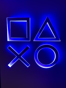 Illuminated geometric shapes including a square, triangle, cross, and circle, glowing with a blue light against a dark background.
