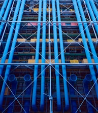A facade with prominent blue industrial pipes running vertically, intersected by smaller pipes and beams. Various structural elements like scaffolding and platforms are present, along with a mix of colors including yellows and greens.