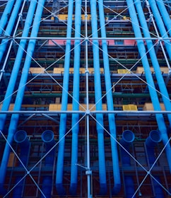 A facade with prominent blue industrial pipes running vertically, intersected by smaller pipes and beams. Various structural elements like scaffolding and platforms are present, along with a mix of colors including yellows and greens.