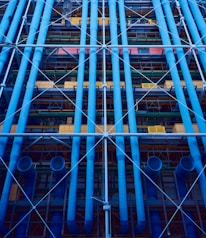 A facade with prominent blue industrial pipes running vertically, intersected by smaller pipes and beams. Various structural elements like scaffolding and platforms are present, along with a mix of colors including yellows and greens.