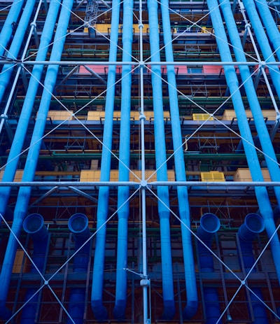 A facade with prominent blue industrial pipes running vertically, intersected by smaller pipes and beams. Various structural elements like scaffolding and platforms are present, along with a mix of colors including yellows and greens.