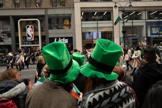A crowd gathers on a city street, watching a parade with participants dressed in festive attire. Several people are wearing large, bright green hats. The background features retail stores and advertisements.