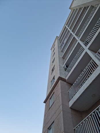 A modern multi-story residential building viewed from an upward angle against a clear blue sky. The facade features multiple balconies with metal railings and a tan exterior.