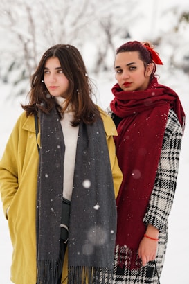 Two women stand side by side in a snowy outdoor setting. They are dressed warmly, with one wearing a yellow coat and a gray scarf, and the other in a checkered coat with a red scarf. Snowflakes are visible in the air, contributing to the wintery atmosphere.