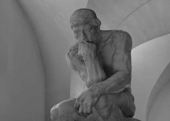 A marble sculpture of a seated man in a contemplative pose, resting his chin on his hand with an intense expression. The setting is minimalist with soft, curved architectural features in the background.