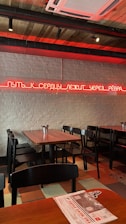 A modern interior of a restaurant or café with wooden tables and black chairs. The walls are made of white brick, and there is a neon sign in red with text written in Russian. The ceiling features exposed ductwork and wooden panels. The room is primarily lit by the red neon sign on the wall.