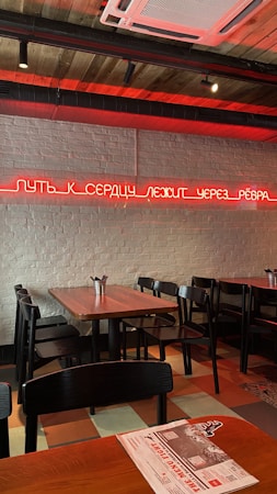 A modern interior of a restaurant or caf&eacute; with wooden tables and black chairs. The walls are made of white brick, and there is a neon sign in red with text written in Russian. The ceiling features exposed ductwork and wooden panels. The room is primarily lit by the red neon sign on the wall.