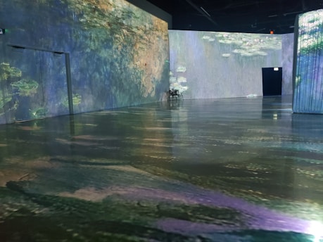 A room with large, immersive projections on the walls depicting Monet's water lilies. The floor reflects the images, creating an illusion of walking on water. Soft lighting enhances the dreamy atmosphere.