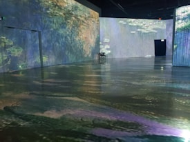 A room with large, immersive projections on the walls depicting Monet's water lilies. The floor reflects the images, creating an illusion of walking on water. Soft lighting enhances the dreamy atmosphere.