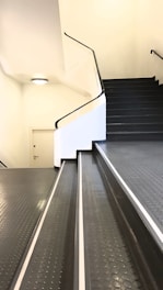 A staircase with an adjoining ramp leading upwards. The surfaces are covered in dark, textured rubber with a dotted pattern. The walls are white and there's a small, round light fixture on the ceiling. A closed door is situated on the landing.