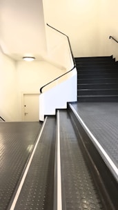 A staircase with an adjoining ramp leading upwards. The surfaces are covered in dark, textured rubber with a dotted pattern. The walls are white and there's a small, round light fixture on the ceiling. A closed door is situated on the landing.
