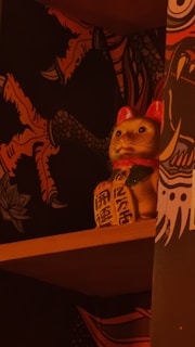 A maneki-neko, or 'beckoning cat' statue sits on a wooden shelf. The background features a dark patterned wallpaper with vibrant, stylized illustrations resembling mythical creatures and nature motifs. The statue is gold with a red collar and raised paw, a common symbol of luck and prosperity in Japanese culture.