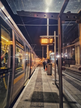 A nighttime scene at a tram station with a well-lit platform and a tram alongside the platform. A few people are present, some waiting, while others are walking. The platform is illuminated by overhead lights, and an electronic display board shows upcoming tram destinations.