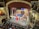 A vibrant theater stage with a group of performers dressed in costumes, holding a large flag, possibly from a staged performance. An orchestra is visible in the pit below the stage. The theater's interior is ornate, with elaborate decorations and balconies.