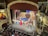 A vibrant theater stage with a group of performers dressed in costumes, holding a large flag, possibly from a staged performance. An orchestra is visible in the pit below the stage. The theater's interior is ornate, with elaborate decorations and balconies.